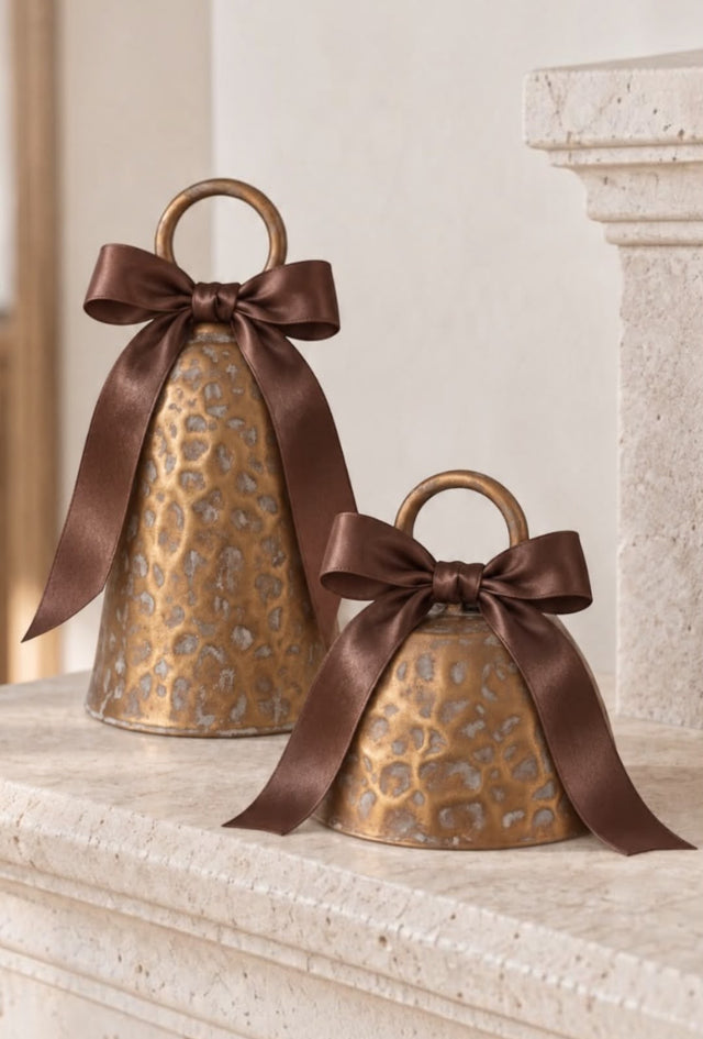 Two hammered antique-gold Christmas bell decorations with brown satin bows on a pale stone mantel