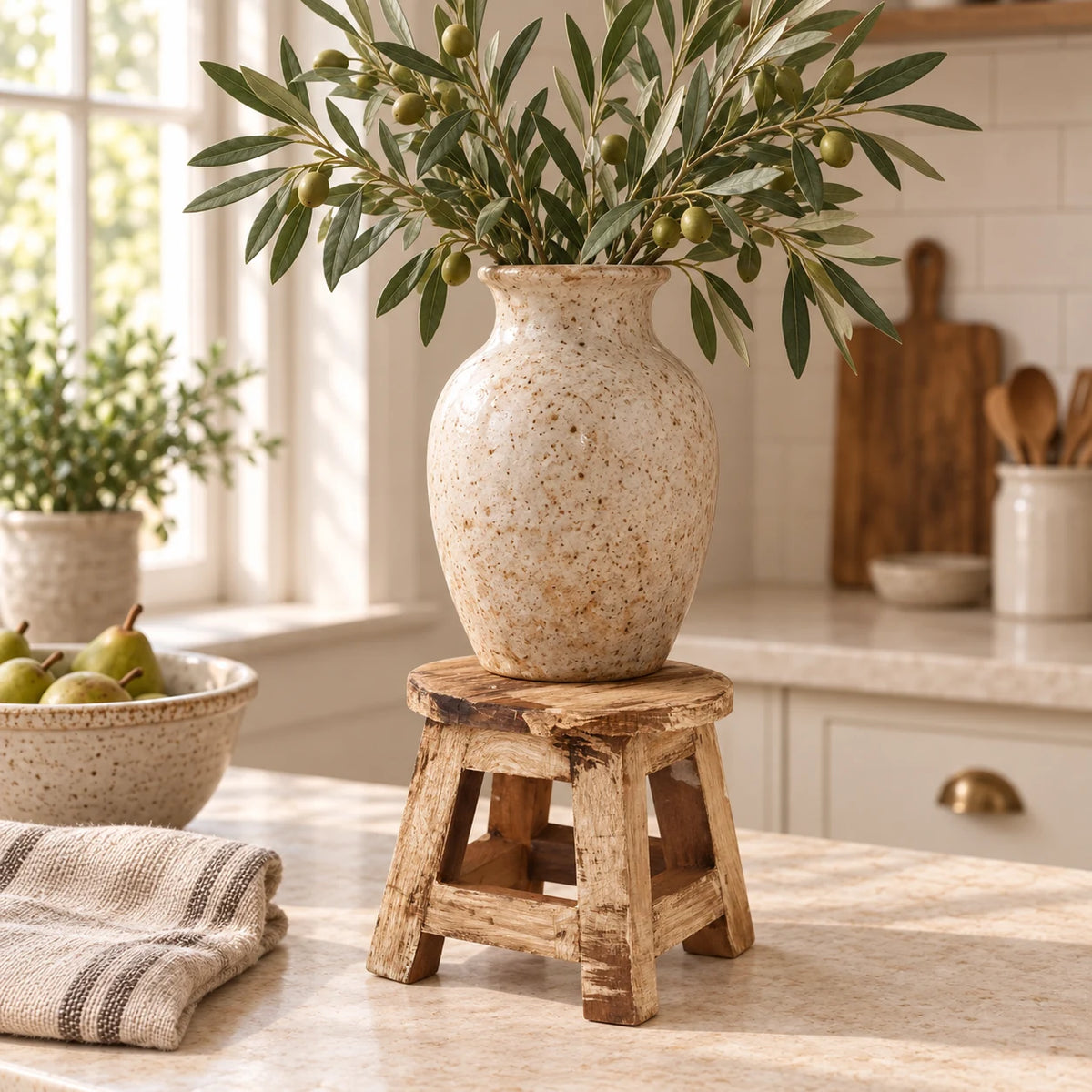 Natural pine mini stool used as a kitchen display riser beneath a large cream ceramic vase of olive foliage.