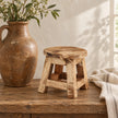 Natural pine mini stool with an empty round top, styled beside a tall brown ceramic jug on a wooden console.