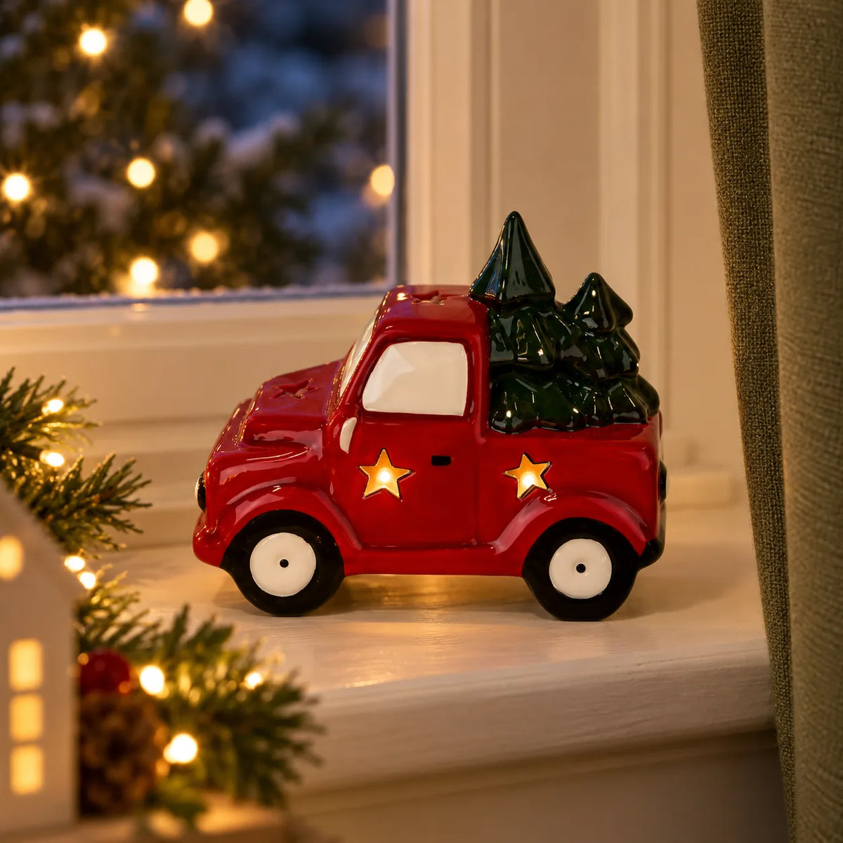 Red ceramic LED truck with two green trees and softly glowing stars on a Christmas windowsill