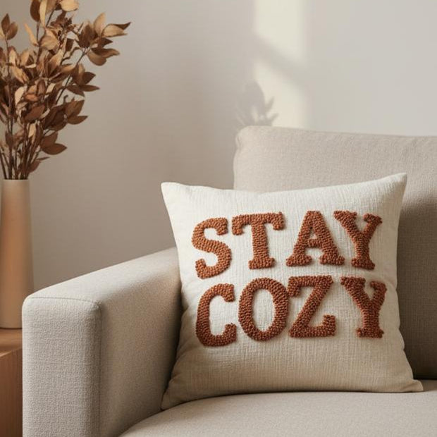 Stay Cozy Rusty Orange Cushion Cover, Stay Cozy cushion on a cream armchair beside autumn foliage