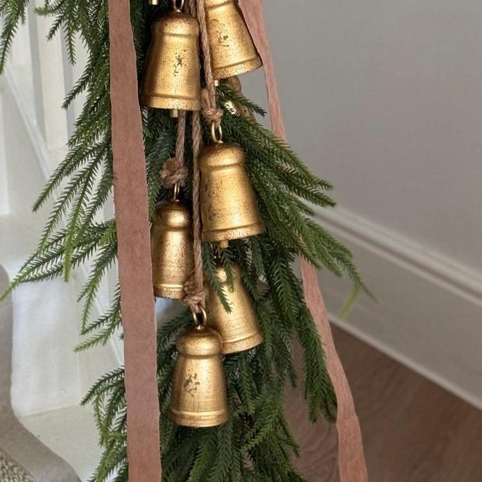 A close-up of a decorative garland with hanging gold bell ornaments, green pine branches, and a dusty pink ribbon in an indoor setting.