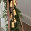 A close-up of a decorative garland with hanging gold bell ornaments, green pine branches, and a dusty pink ribbon in an indoor setting.