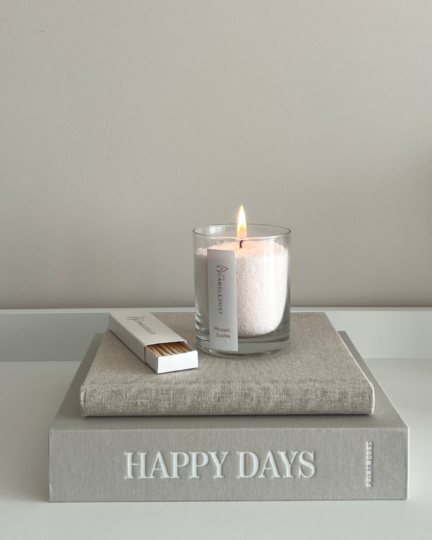 Candle in a glass holder on a book titled 'Happy Days' with a matchbox on a light gray background