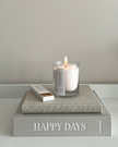 Candle in a glass holder on a book titled 'Happy Days' with a matchbox on a light gray background