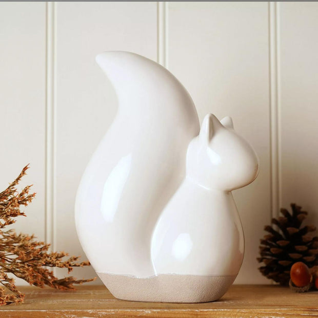 A glossy white ceramic squirrel figurine displayed on a wooden shelf against a cream paneled wall, with dried branches and a pinecone nearby.