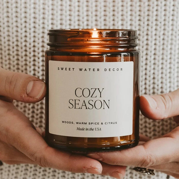 Hands holding a lit Sweet Water Decor Cozy Season 9 oz candle in an amber jar against a cream knitted jumper.