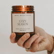Lit Cozy Season 9 oz candle in an amber jar balanced on one hand, with its cream fragrance label facing forwards.