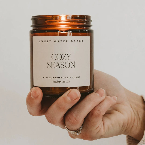 Lit Cozy Season 9 oz candle in an amber jar balanced on one hand, with its cream fragrance label facing forwards.