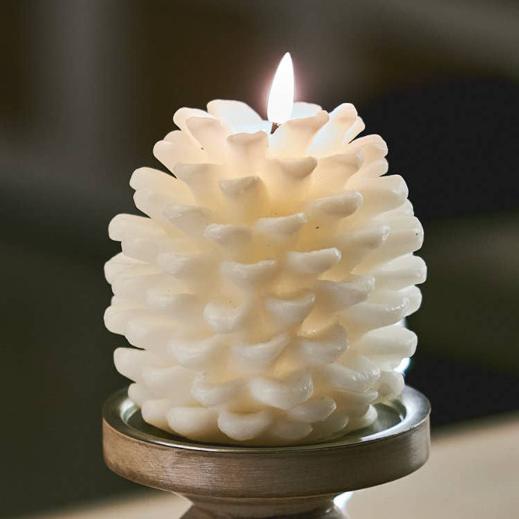A white pinecone-shaped candle with a lit flame, displayed on a round wooden holder against a softly blurred indoor background.