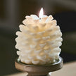 A white pinecone-shaped candle with a lit flame, displayed on a round wooden holder against a softly blurred indoor background.