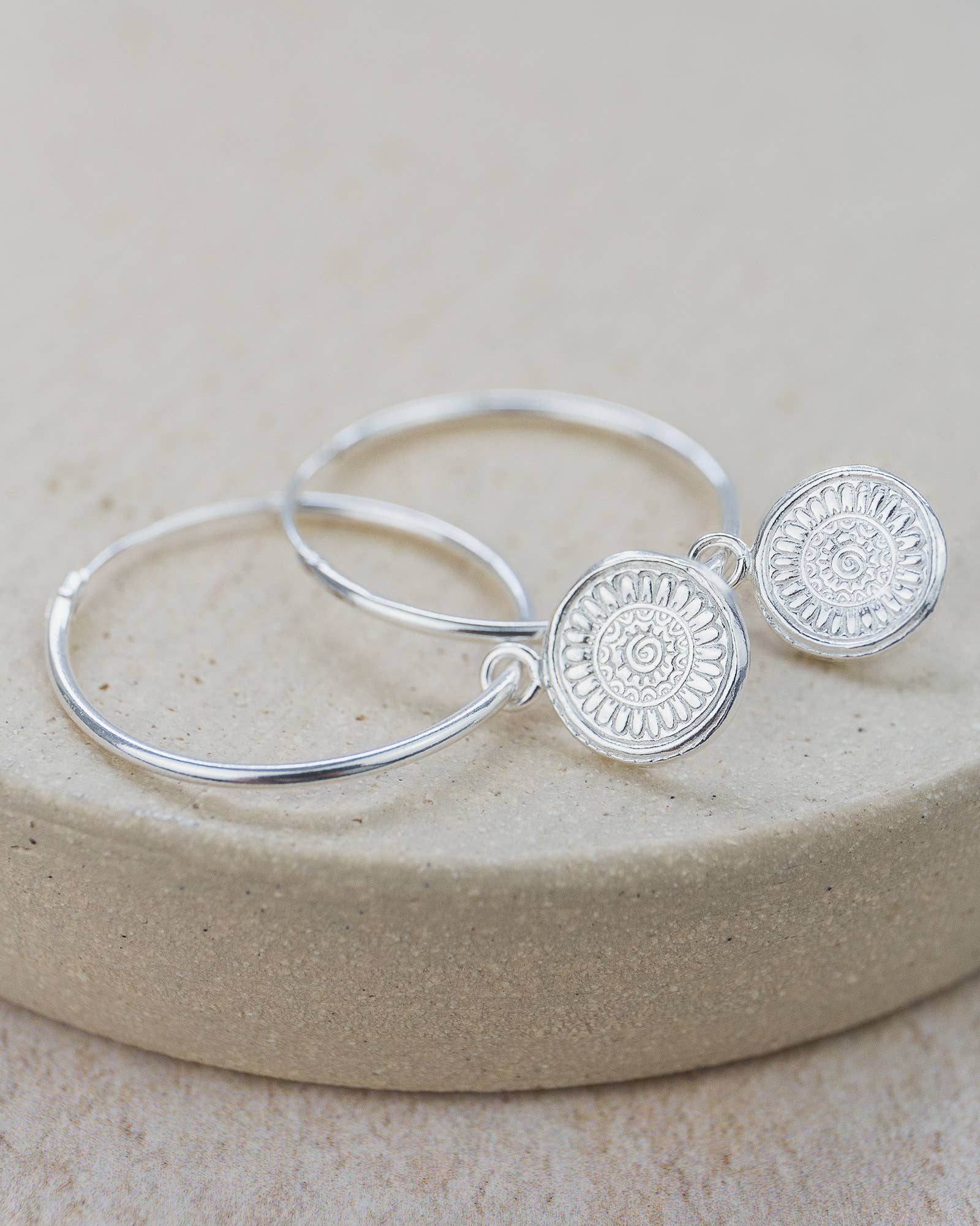 Silver hoop earrings with circular designs on a beige stone surface