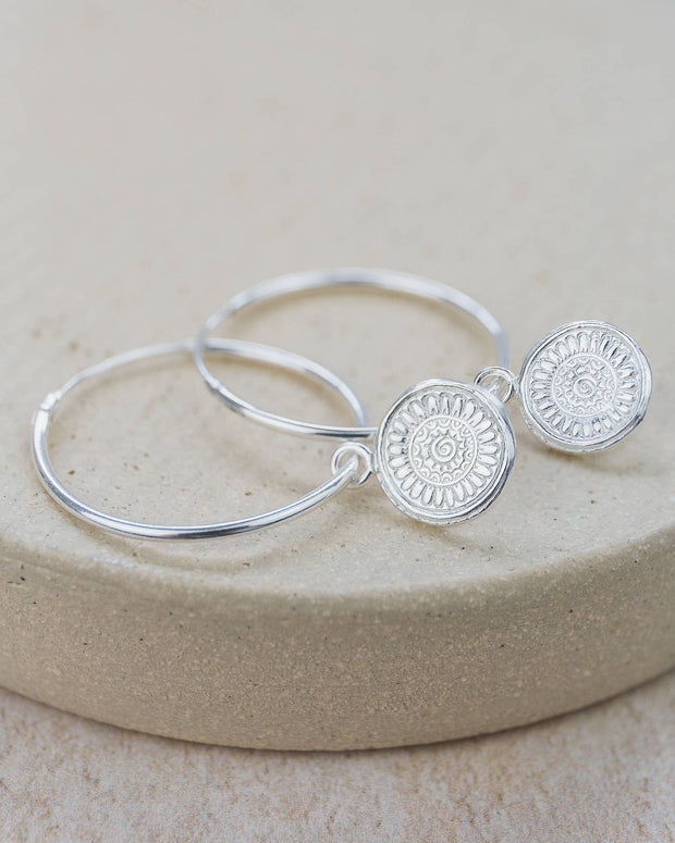 Silver hoop earrings with circular designs on a beige stone surface