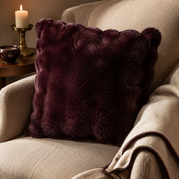 Burgundy Luxury Faux Fur Cushion, burgundy faux fur cushion on a cream armchair beside a candle
