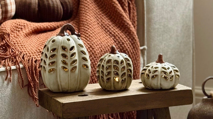 A styled autumn living room corner with a light gray sofa draped in a rust-orange throw blanket, decorative pumpkins on a small wooden stool, and rustic metal bells on a light wood floor.