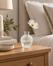Glass acorn vase with a white flower by a bed