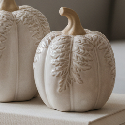 Two ribbed ceramic pumpkins in a warm off-white finish with embossed leaf patterns, displayed on a stack of books in a cozy, softly lit home interior.