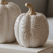 Two ribbed ceramic pumpkins in a warm off-white finish with embossed leaf patterns, displayed on a stack of books in a cozy, softly lit home interior.