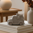 A small gray decorative figurine shaped like a vintage pickup truck carrying logs, displayed on top of stacked books in a softly lit interior setting.