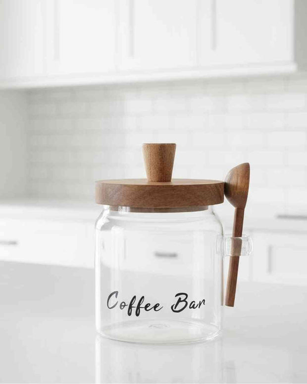 Clear glass coffee storage jar with dark wood lid and spoon, ideal for kitchen and dining organization.