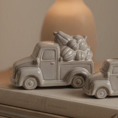 Two silver-toned vintage pickup truck figurines carrying bundles of Christmas trees, displayed on a light gray surface against a white paneled background.