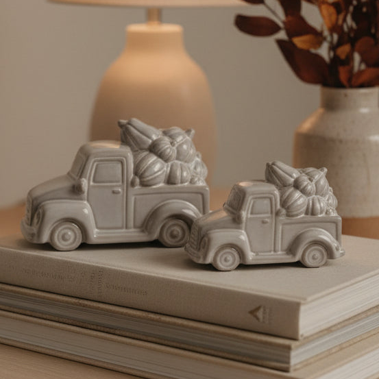 Two small gray ceramic pickup trucks carrying pumpkins, displayed on stacked books with a softly blurred lamp and vase in the background.
