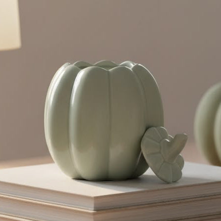 A small matte sage green decorative pumpkin sculpture with a matching leaf detail, displayed on top of a stack of books on a wooden table in a softly lit interior setting.