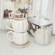 A stack of two ribbed ceramic mugs in warm off-white sits on a woven coaster next to a matching ribbed decorative pumpkin, with an open magazine in the background showing a dining room scene.