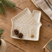 Cream ceramic gingerbread house trinket plate lying flat with pine cones and a small brass trinket