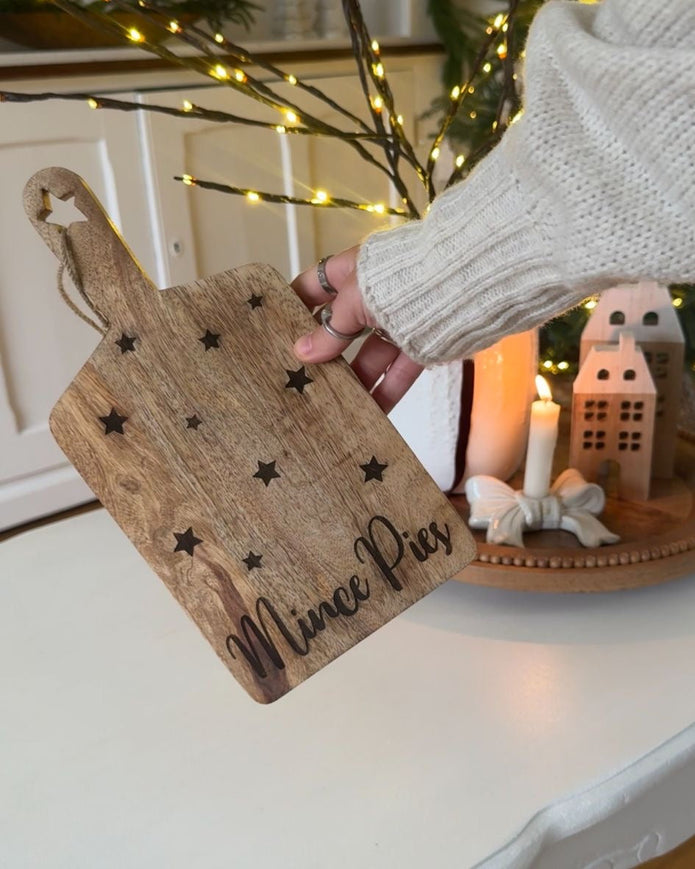 Person holding a wooden cutting board with 'Mince Pies' written on it in a festive setting.
