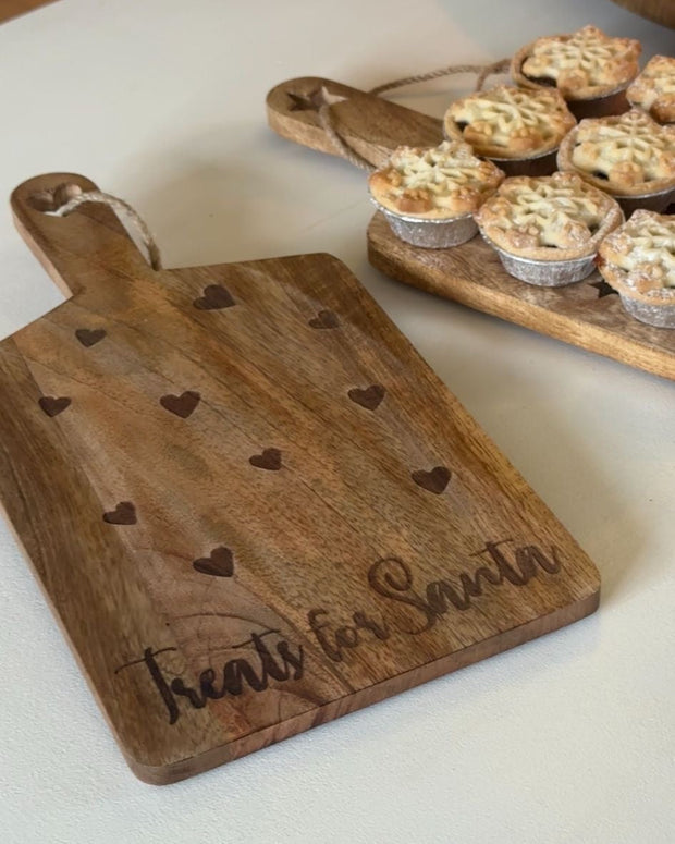 Wooden cutting board with heart designs and 'Santa's Little Helper' engraving on a white surface.
