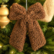 A brown curly-textured bow Christmas ornament hanging on a green Christmas tree with warm lights.