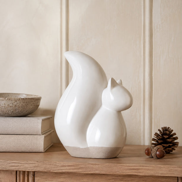 A glossy white ceramic squirrel figurine with a light beige base, displayed on a wooden shelf next to a pinecone and books.