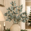 Artificial eucalyptus branch with muted blue-green leaves and small dark berries, arranged in a textured cream ceramic vase on a tabletop in a warmly lit indoor setting.