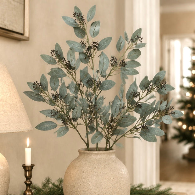 Artificial eucalyptus branch with muted blue-green leaves and small dark berries, arranged in a textured cream ceramic vase on a tabletop in a warmly lit indoor setting.