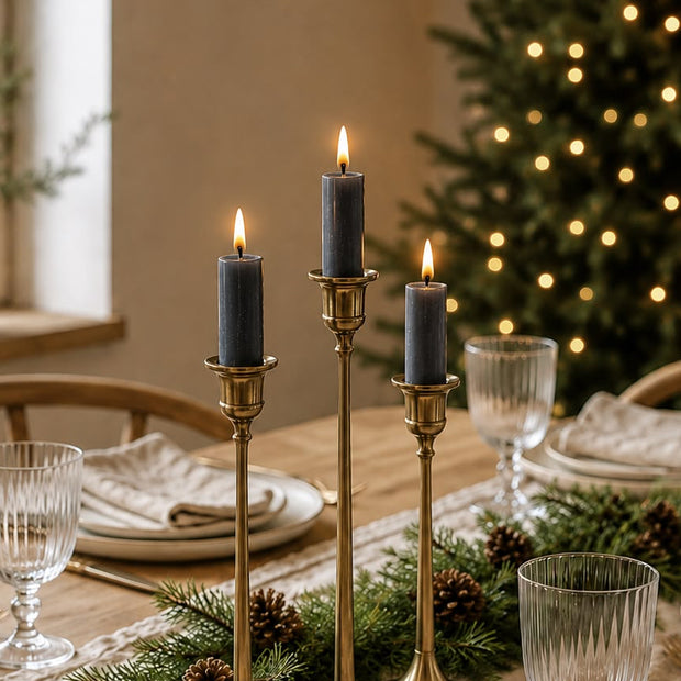 Three brass candlesticks with lit blue-gray taper candles are arranged on a dining table with plates, glassware, pine branches, and pinecones, with a softly blurred Christmas tree in the background.