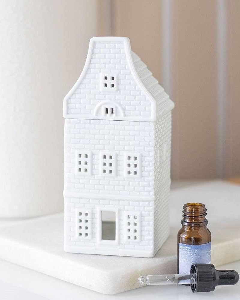 White ceramic house-shaped diffuser on a white surface with a dropper bottle and small container.