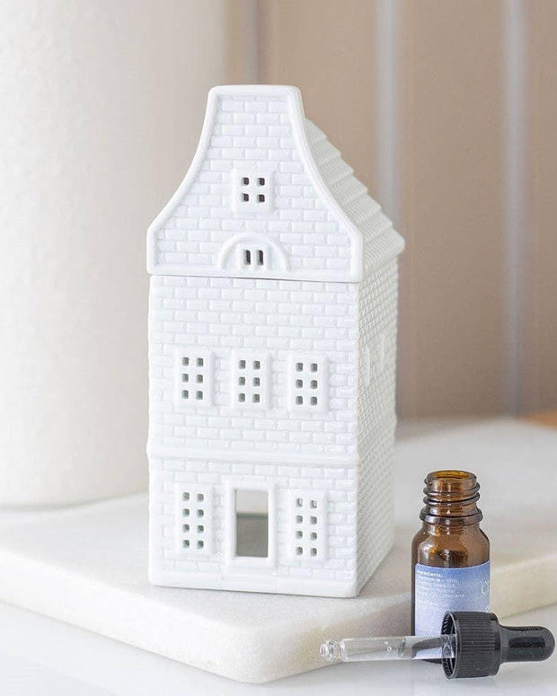 White ceramic house-shaped diffuser on a white surface with a dropper bottle and small container.