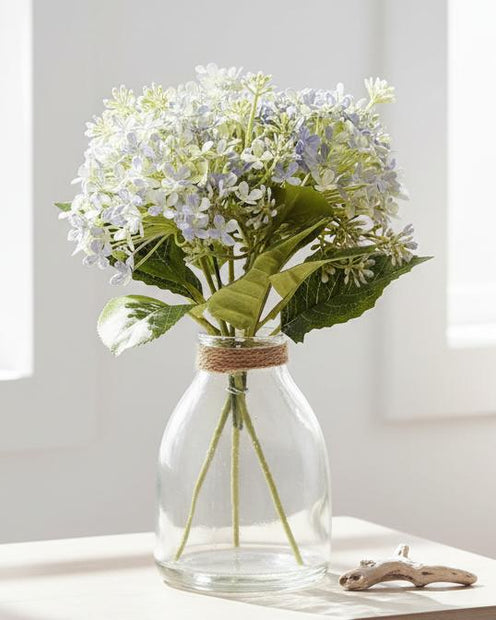 Pale blue artificial flower arrangement in a glass vase by Heaven Sends, showcasing delicate blooms and greenery.