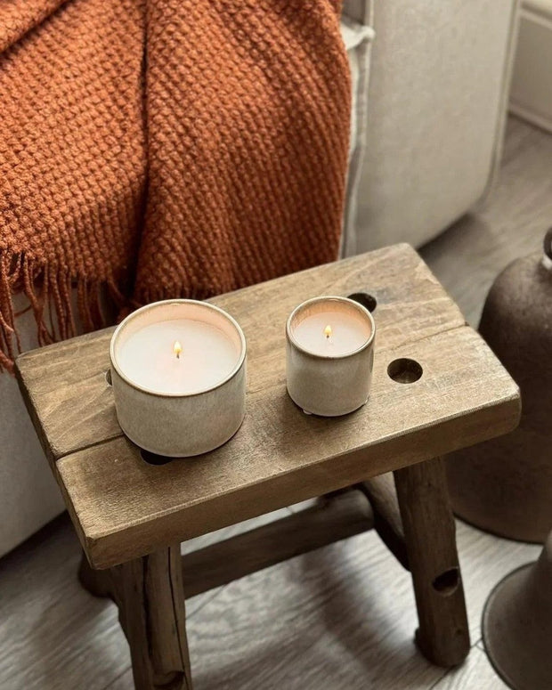 Two candles on a small wooden stool with a cozy blanket and chair in the background.