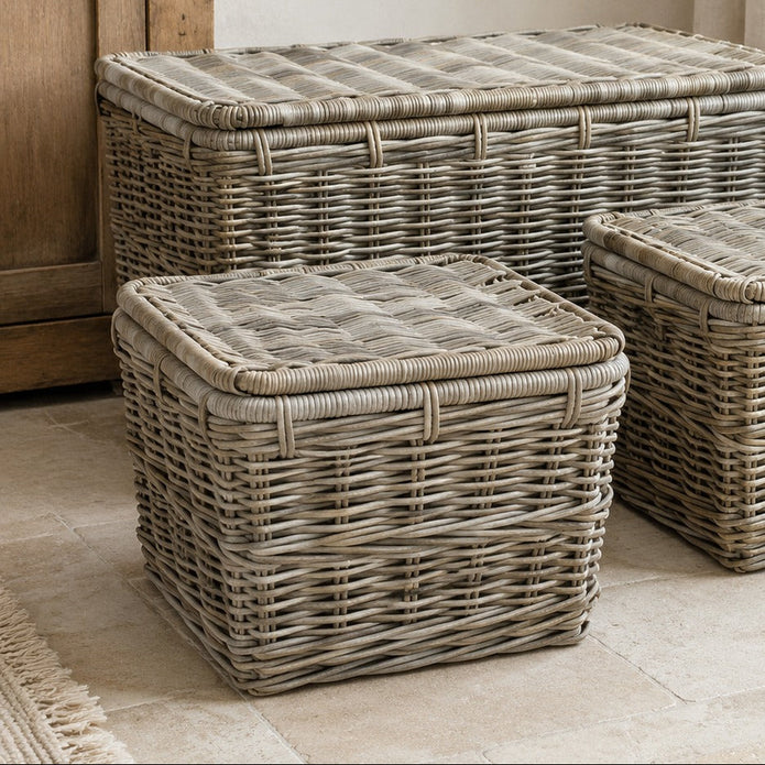 Three wicker storage bins in different sizes arranged on a light stone floor in a softly styled room with a wooden cabinet, curtain, and potted plant.