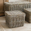 Three wicker storage bins in different sizes arranged on a light stone floor in a softly styled room with a wooden cabinet, curtain, and potted plant.
