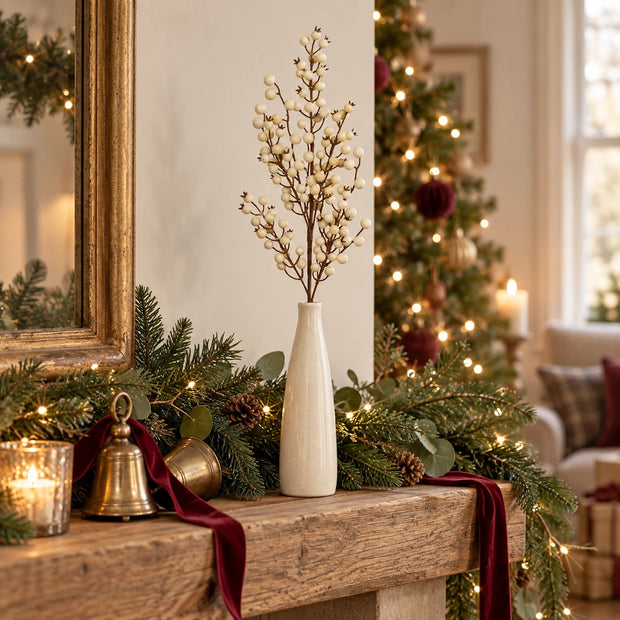 Cream berry stem in a vase on a Christmas mantel with greenery, burgundy ribbon and brass bells.