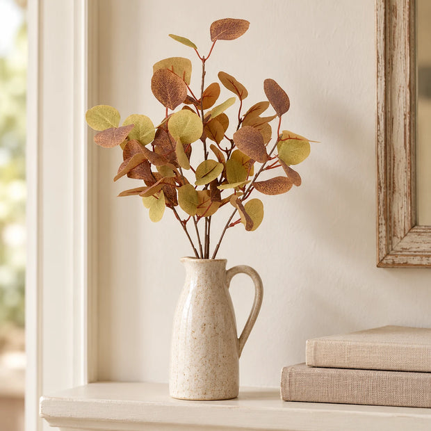 Autumn eucalyptus sprig in a stoneware jug on a cream shelf.