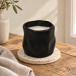 Unlit black vessel candle with ivory wax on a stone coaster and oak side table