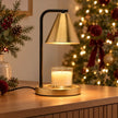 Brass-effect candle warmer lamp with black stem warming an unlit jar candle beside Christmas lights