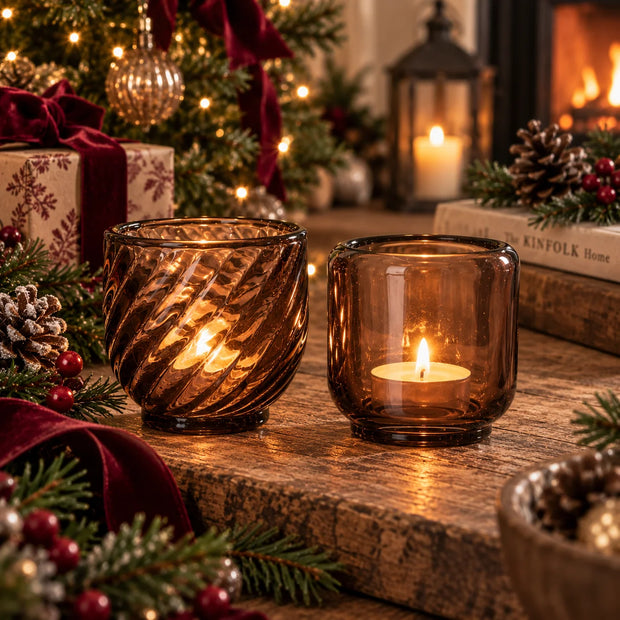 Patterned and plain brown glass tealight candle holders glowing beside Christmas greenery and burgundy ribbon