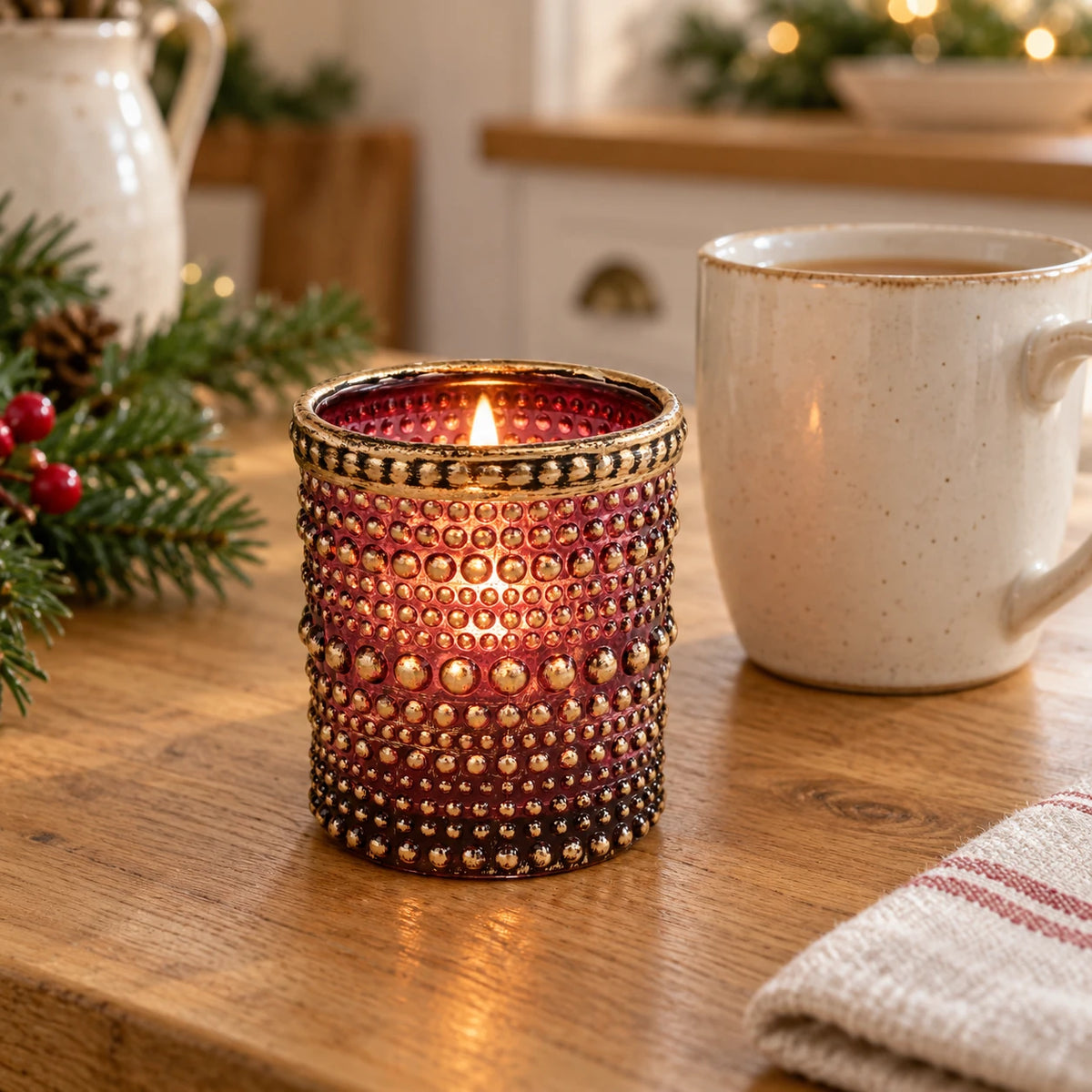 Burgundy dotted glass tealight holder with distressed gold rim, beside a mug of tea on a kitchen counter, showing its compact size.