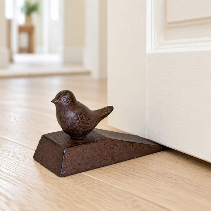 Rust-brown cast iron bird door wedge, 12cm wide, shown beneath a cream door on an oak floor.