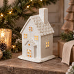 Large cream ceramic house tealight holder glowing beside Christmas greenery and pinecones